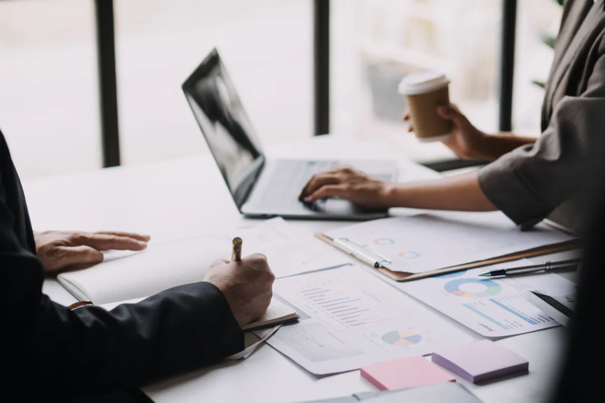 Duas pessoas em uma reunião de negócios, com gráficos de desempenho e laptop sobre a mesa, enquanto uma delas segura um copo de café.