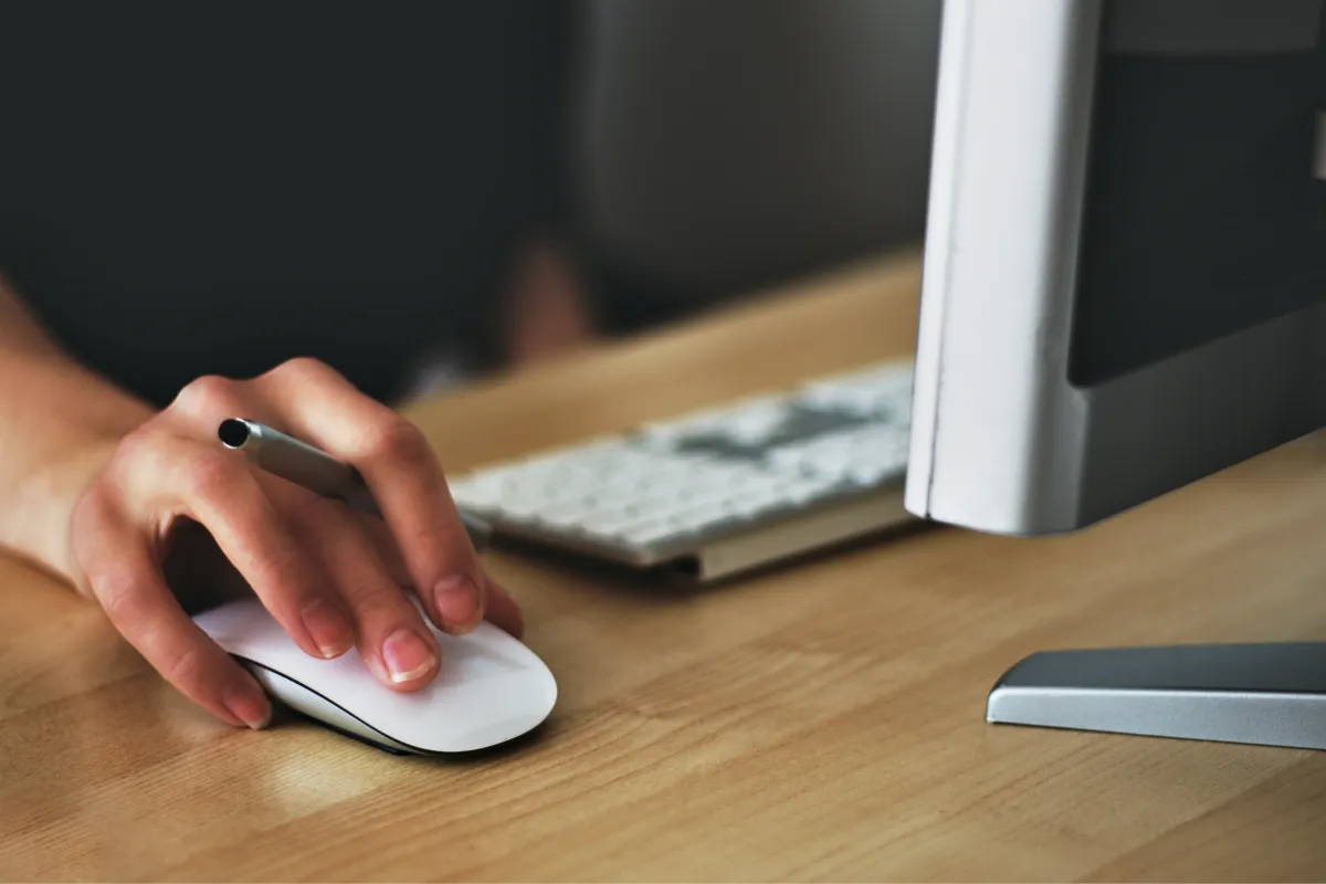 Mão segurando um mouse branco sobre uma mesa de madeira clara, com teclado e monitor ao fundo, representando uma rotina de trabalho no computador.
