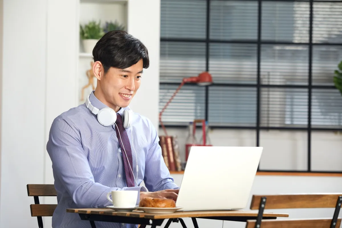 Homem asiático sorridente usando camisa social e fones de ouvido ao pescoço, digitando em notebook branco em uma cafeteria moderna com xícara de café e croissant na mesa