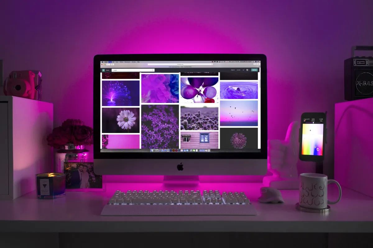 Ambiente de trabalho moderno iluminado por luzes roxas, com um computador Apple exibindo imagens de flores e objetos em tons de roxo. Um celular e uma caneca complementam o cenário.