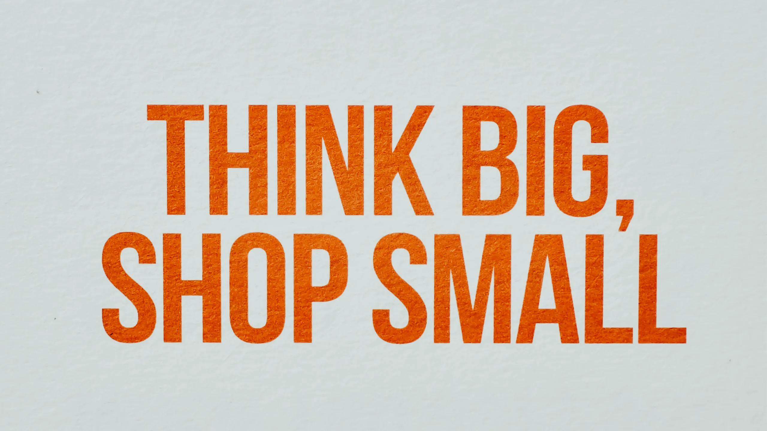 Frase ‘THINK BIG, SHOP SMALL’ em letras maiúsculas laranja sobre fundo claro.