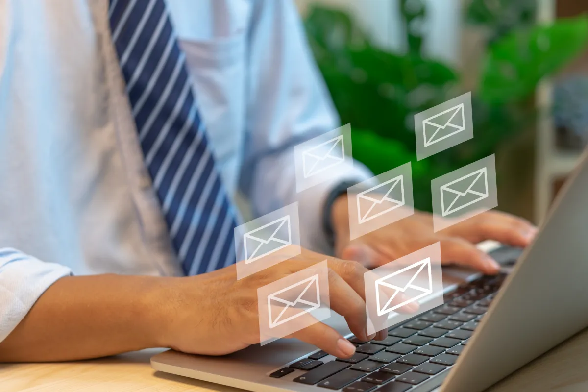 Mãos de pessoa vestindo camisa social e gravata digitando em notebook, com ícones de envelope semitransparentes flutuando em primeiro plano
