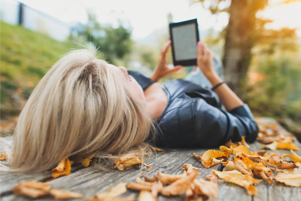 Mulher deitada sobre folhas secas lendo em um e-reader em dia de outono.