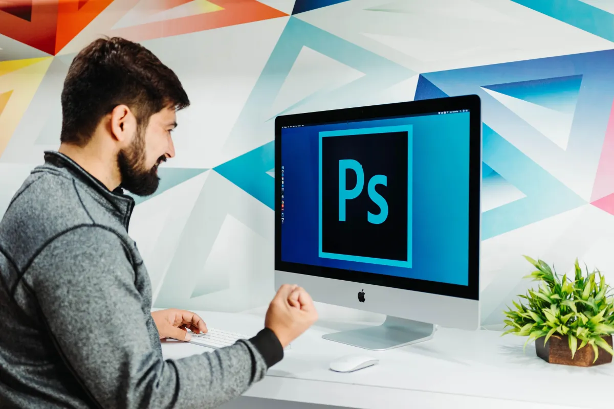 Homem sorridente sentado em frente a um iMac com a tela exibindo o logotipo do Photoshop, em ambiente moderno com parede colorida e planta decorativa