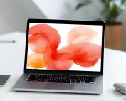 Notebook moderno em uma mesa branca, mostrando na tela um fundo abstrato em aquarela nas cores vermelho e laranja, com objetos de escritório ao redor.