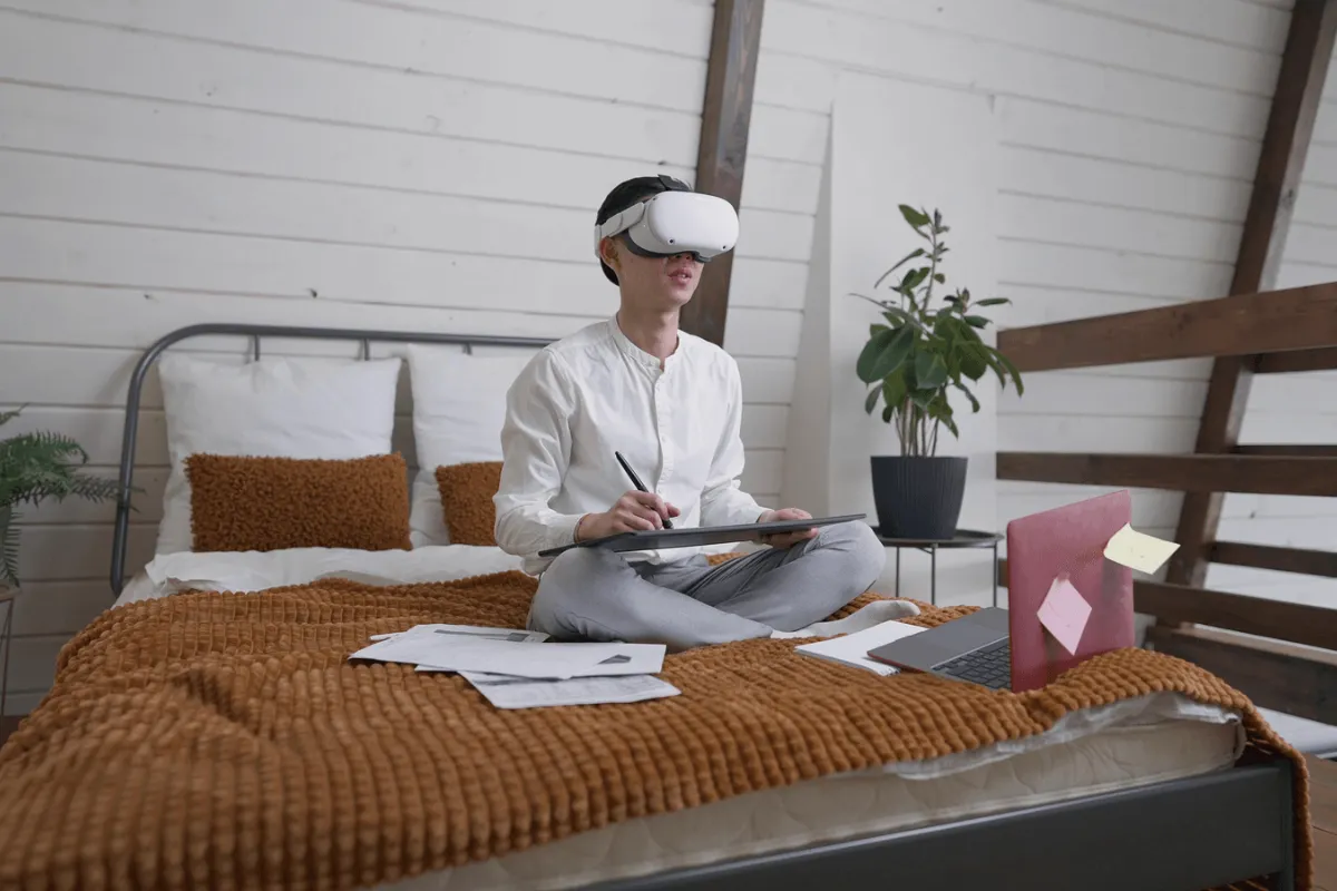 Pessoa sentada em cama com notebook, papéis e tablet, utilizando óculos de realidade virtual para estudo ou trabalho remoto.