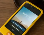 Smartphone com capa amarela exibindo a tela inicial do Instagram, com opções de login e registro via Facebook ou e-mail, apoiado sobre mesa de madeira.