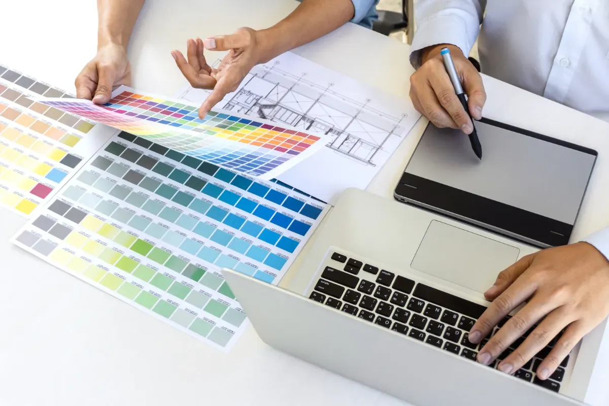 Duas pessoas analisando paleta de cores impressa ao lado de um laptop e mesa digitalizadora durante processo de design gráfico.